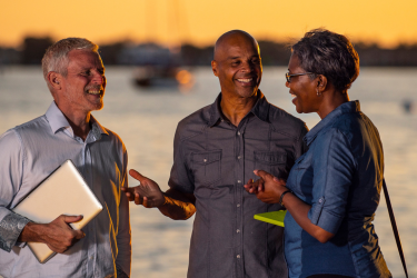 Three people talk near the marina with sailboat in the background