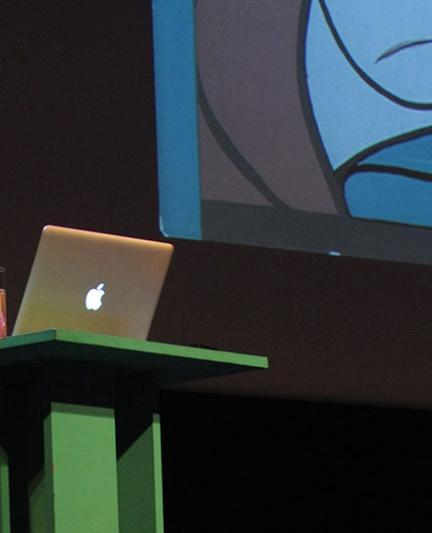 A female speaker is presenting at the podium, operating her macbook.