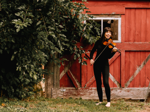 Violinist Tessa Lark posing with violin outside in grass in front of red barn doors. 