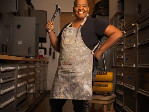 Artist Kendra Frorup stands with apron on and hammer in hand.