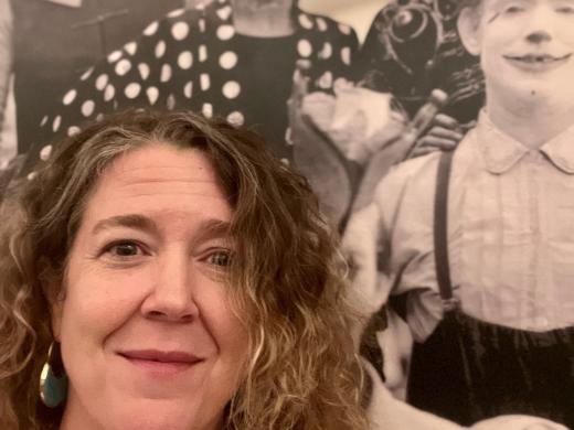 Headshot of Jennifer Lemmer Posey with long hair and smile, plus circus poster behind her.