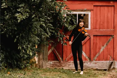 Violinist Tessa Lark posing with violin outside in grass in front of red barn doors. 