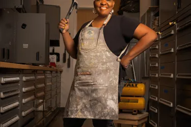 Artist Kendra Frorup stands with apron on and hammer in hand.