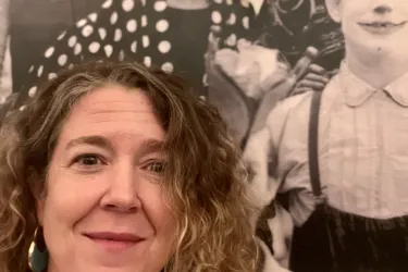 Headshot of Jennifer Lemmer Posey with long hair and smile, plus circus poster behind her.